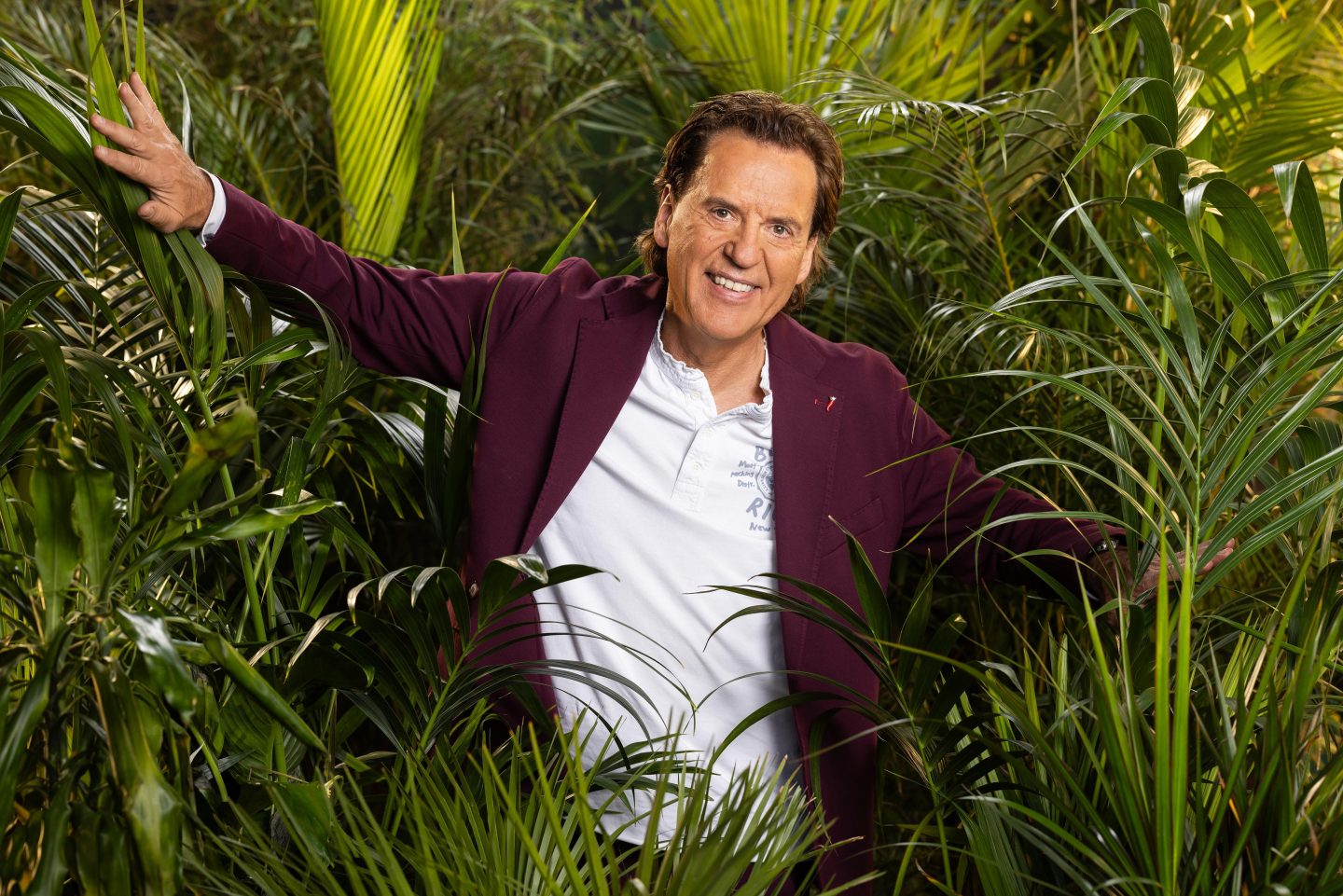 Jörg Dahlmann, lächelnd in einem weißen Polo-Shirt und bordeauxfarbenen Blazer, mit ausgebreiteten Armen zwischen tropischen Pflanzen im Dschungel.







