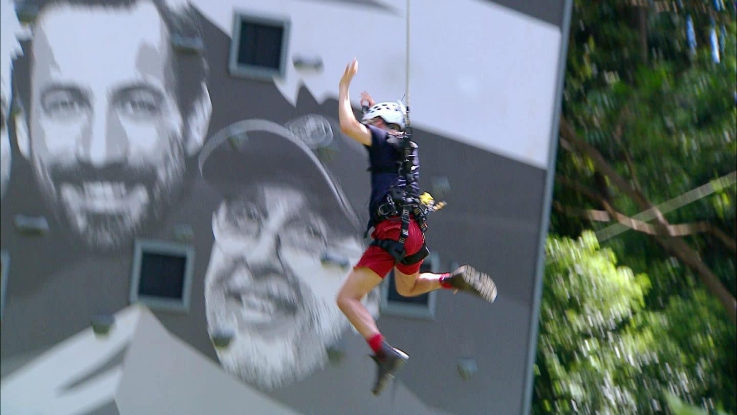 Sam Dylan fällt bei der Dschungelprüfung „Karriere-Leider zum Mount IBES“ von der Kletterwand in die Tiefe. Er trägt ein Sicherheitsgeschirr und Helm, während er mit ausgestrecktem Arm rückwärts stürzt. Im Hintergrund sind die großformatigen Porträts von Sonja Zietlow, Jan Köppen und einem weiteren Gesicht auf der Kletterwand zu sehen. Trotz Höhenangst hat Sam sich der Herausforderung gestellt, doch seine Kräfte verlassen ihn an der Wand, sodass er loslassen muss.
