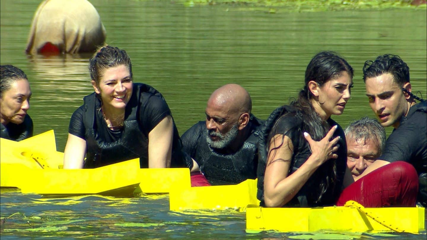 Die Dschungelcamp-Bewohner Lilly Becker, Nina Bott, Pierre Sanoussi-Bliss, Yeliz Koç, Jürgen Hingsen und Timur Ülker während einer Wasserprüfung. Sie tragen schwarze Westen und klammern sich an gelbe Schwimmhilfen, während sie sichtlich erschöpft im Wasser treiben.