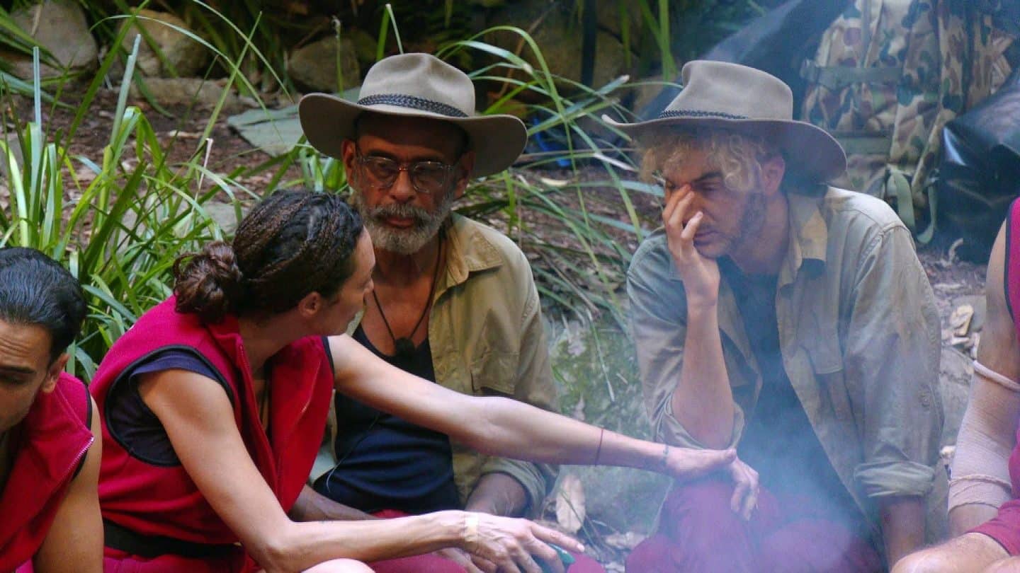 Sam Dylan (rechts) sitzt erschöpft am Lagerfeuer im Dschungelcamp, stützt seinen Kopf auf die Hand und wirkt nachdenklich. Er wurde zu seiner insgesamt achten Dschungelprüfung gewählt. Lilly Becker (links) lehnt sich zu ihm und scheint ihn zu motivieren, während Pierre Sanoussi-Bliss (Mitte) mit neutralem Blick zuhört. Alle drei tragen typische Dschungelcamp-Kleidung, während im Hintergrund Pflanzen und Campingausrüstung zu sehen sind.