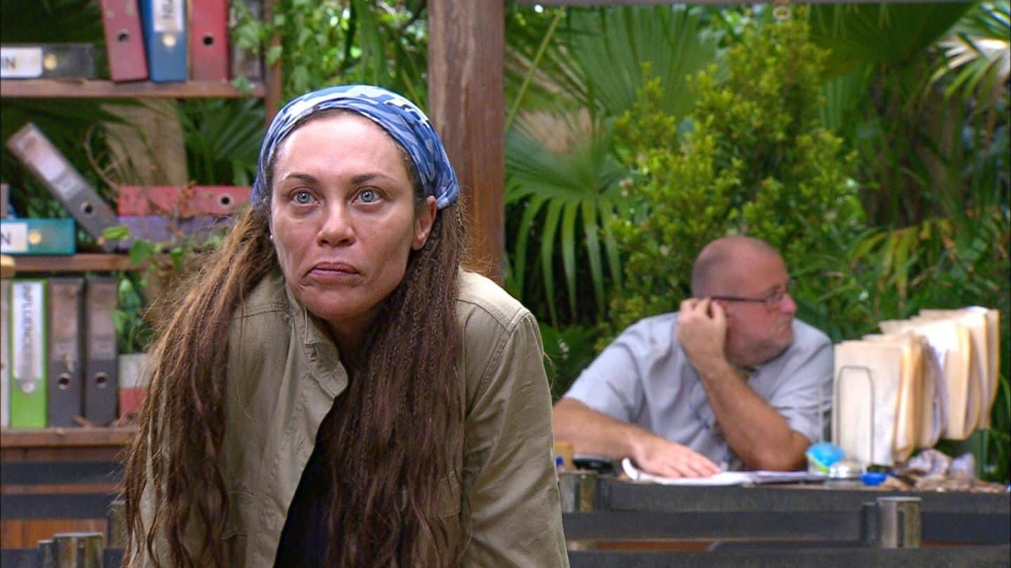 Lilly Becker in der Dschungelprüfung „Bährufs-Bähratung“, trägt eine beige Jacke und ein blaues Kopftuch, während sie mit angespannter Miene in die Kamera blickt. Im Hintergrund sitzt ein Mann an einem Schreibtisch mit Akten.