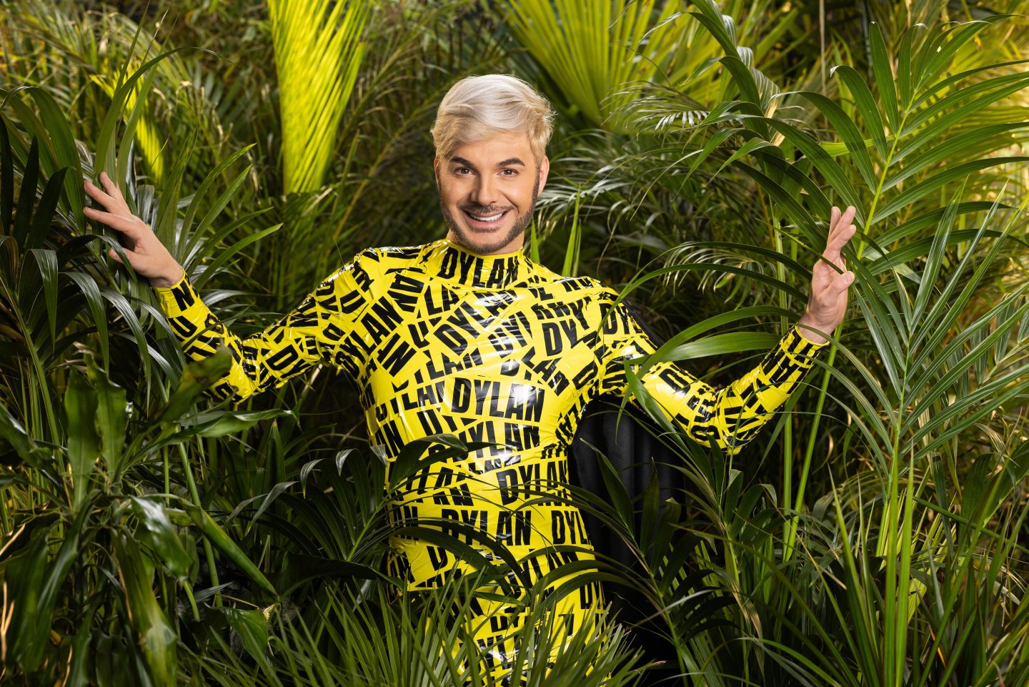 Sam Dylan, in einem auffälligen gelben Latexanzug mit seinem Namen als schwarzem Schriftzug bedruckt, lächelnd zwischen tropischen Pflanzen im Dschungel.







