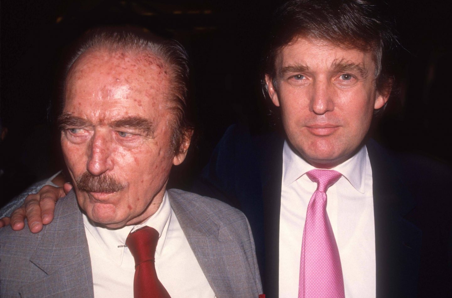 Donald Trump (rechts) mit seinem Vater Fred Trump (links) bei einer gemeinsamen Veranstaltung. Fred Trump trägt einen grauen Anzug mit roter Krawatte, während Donald Trump eine rosafarbene Krawatte trägt.