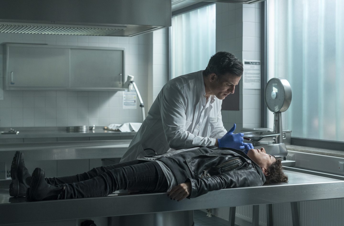 Harry Möller (Maria Ketikidou) liegt regungslos auf einem metallenen Obduktionstisch, gelähmt durch ein Gift. Dr. Torben Achs (Torben Liebrecht) steht über ihr und beugt sich mit einer Spritze in der Hand zu ihr hinunter. Sein ernster Ausdruck und die sterile Umgebung eines Labors oder einer Pathologie unterstreichen die bedrohliche Situation.






