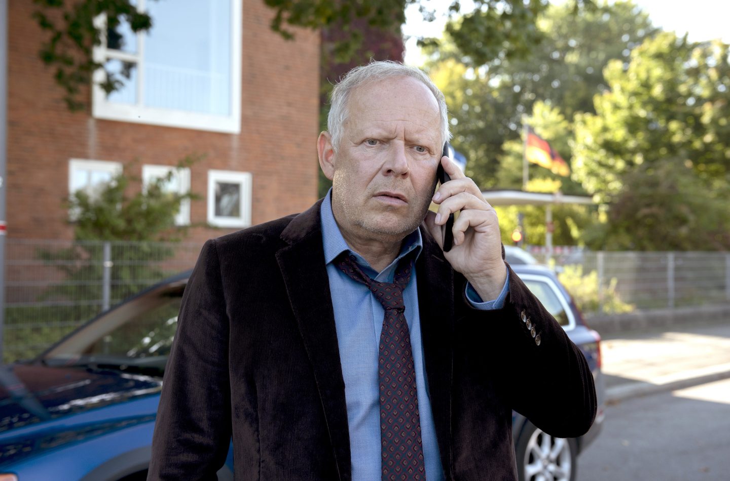 Kommissar Klaus Borowski, gespielt von Axel Milberg, steht draußen vor einem blauen Auto und telefoniert angespannt. Im Hintergrund sind ein rotes Backsteingebäude, Bäume und eine wehende Deutschlandflagge zu sehen. Die Szene vermittelt Dramatik und Konzentration.