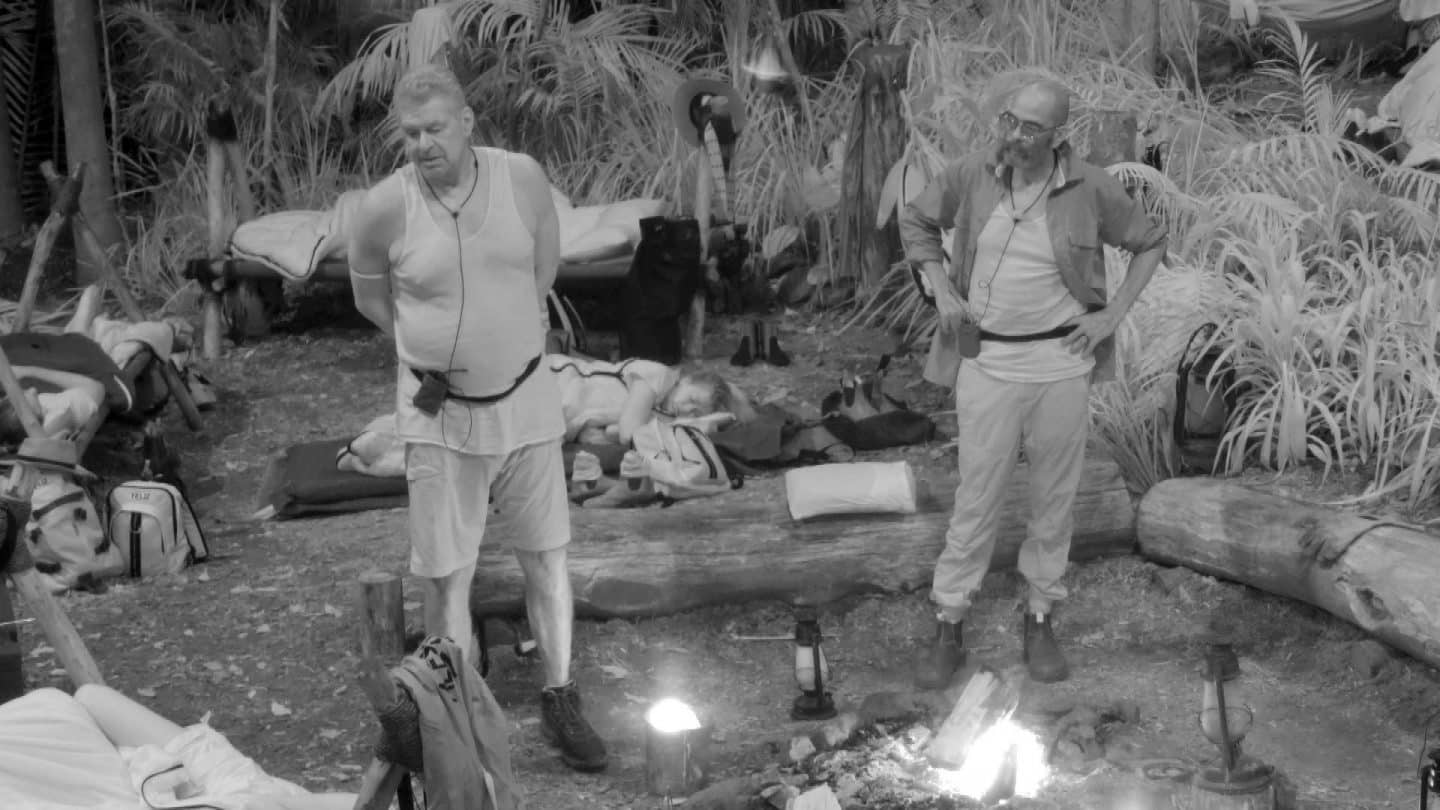 Jürgen Hingsen (links) und Pierre Sanoussi-Bliss (rechts) stehen während ihrer gemeinsamen Nachtwache am Lagerfeuer im Dschungelcamp. Beide tragen helle Kleidung und wirken in ein Gespräch vertieft. Im Hintergrund sind Schlafplätze der anderen Camper zu sehen, während eine Person auf einer Liege ruht. Die Szene ist in Schwarz-Weiß gehalten, was die nächtliche Atmosphäre unterstreicht. Während der Wache nutzt Hingsen die Gelegenheit, um über die anderen Camp-Bewohner zu sprechen.