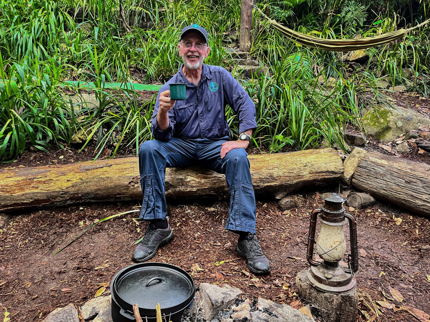 Dr. Bob, der bekannte Arzt des Dschungelcamps, sitzt auf einem Baumstamm im Camp, hält eine Tasse in der Hand und lächelt. Im Hintergrund sind eine Hängematte, Pflanzen und Dschungel-Atmosphäre zu sehen.







