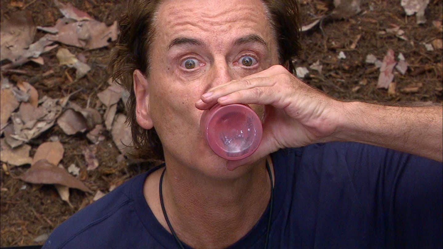 Jörg Dahlmann während der Dschungelprüfung „Bährufs-Bähratung“, trinkt mit weit aufgerissenen Augen eine rosa Flüssigkeit aus einem kleinen Becher, während er einen blauen Pullover trägt.