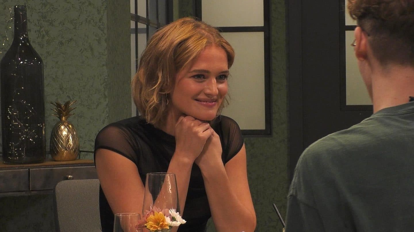 Chiara Moon Horst, eine junge Frau mit kinnlangem, welligem blondem Haar, sitzt an einem Tisch in einem stilvollen Restaurant. Sie trägt ein schwarzes, leicht durchsichtiges Oberteil und lächelt freundlich, während sie ihre Hände unter ihr Kinn stützt. Im Vordergrund ist die Silhouette ihres Gesprächspartners zu sehen, im Hintergrund eine goldene Ananas-Dekoration und stilvolle Beleuchtung.