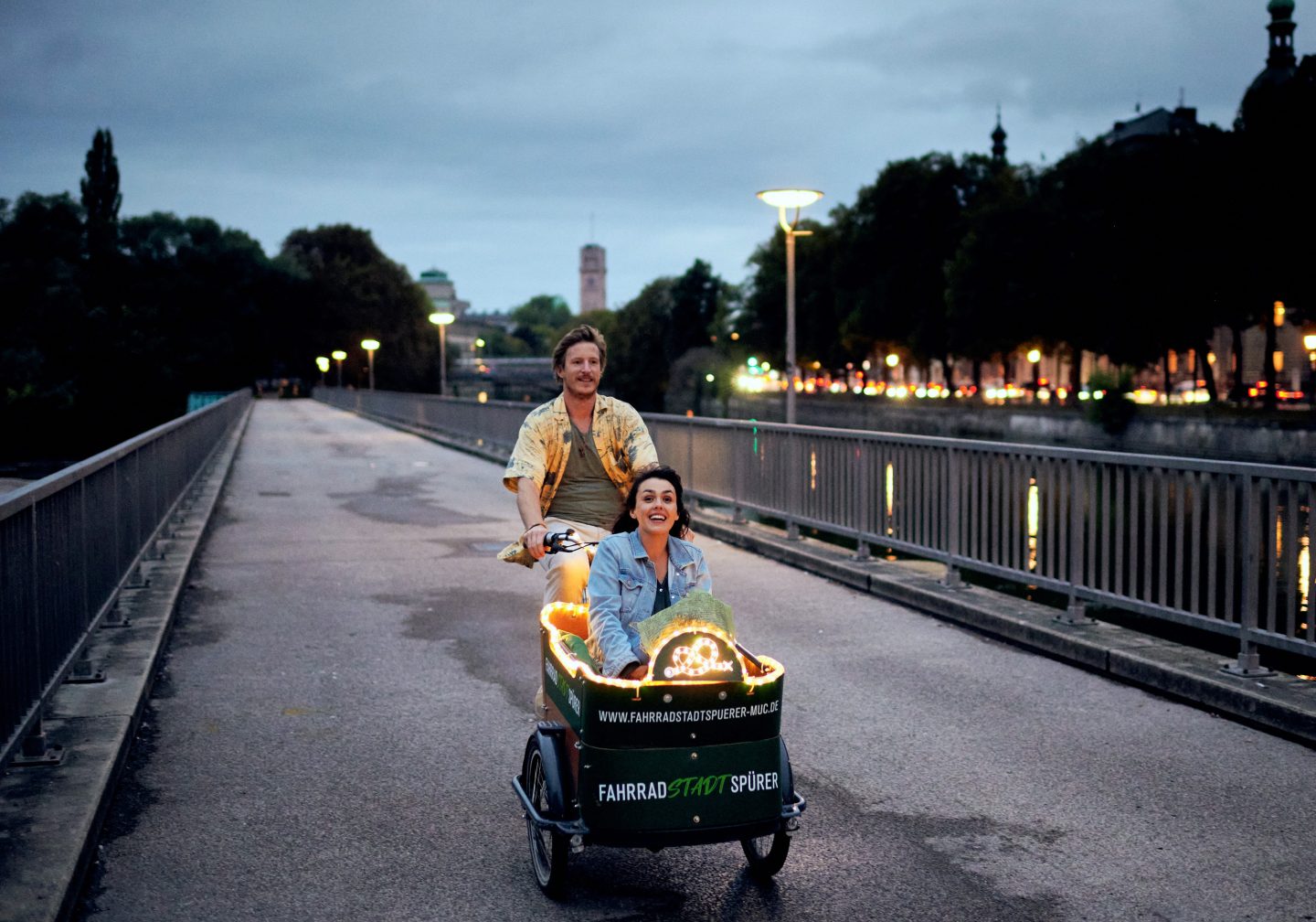 Szene aus „Eine Liebe später“: Ein Mann mit Hawaiihemd (Golo Euler) fährt eine Frau (Lucie Heinze) in einem beleuchteten Lastenfahrrad über eine Brücke in München bei Abenddämmerung. Die Stadtlichter und der Fluss sind im Hintergrund sichtbar.