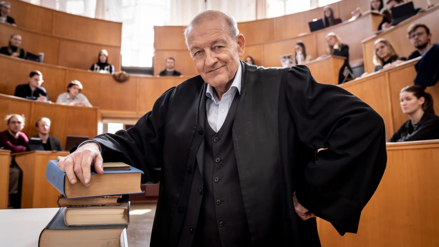 "Wilsberg - Über dem Gesetz": Wilsberg (Leonard Lansink) steht in einem Unihörsaal, sein Arm lehnt auf einem Stapel mit dicken Büchern, er blickt lächelnd in die Kamera. In den Sitzreihen hinter ihm sitzen Studierende und schauen in Richtung Wilsberg.
