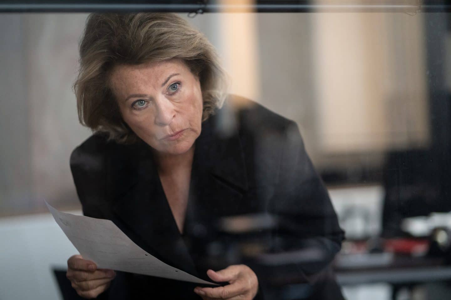 "Wilsberg - Über dem Gesetz": Anna Springer (Rita Russek) blickt unter einer Jalousie ihres Bürofensters hindurch. Sie hält ein gelochtes Blatt Papier in den Händen.
