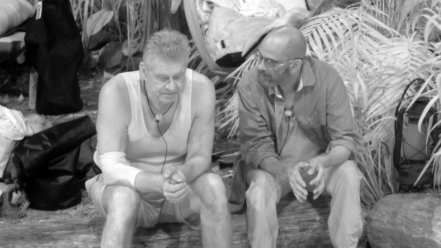 Jürgen Hingsen (links) und Pierre Sanoussi-Bliss (rechts) sitzen während ihrer Nachtwache auf einem Baumstamm im Dschungelcamp. In nachdenklicher Stimmung sprechen sie miteinander, umgeben von Pflanzen und Campingausrüstung. Das Bild ist in Schwarz-Weiß gehalten.