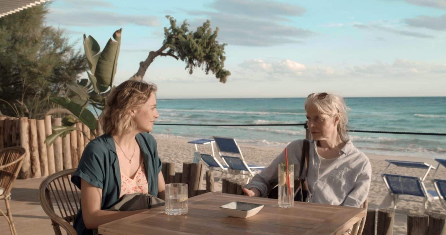 "Ein Sommer in Italien": Billie (Hanna Plaß) und Hella (Corinna Kirchhoff) sitzen an einer Sandstrandbar und schauen einander an. Im Hintergrund liegt das Meer.