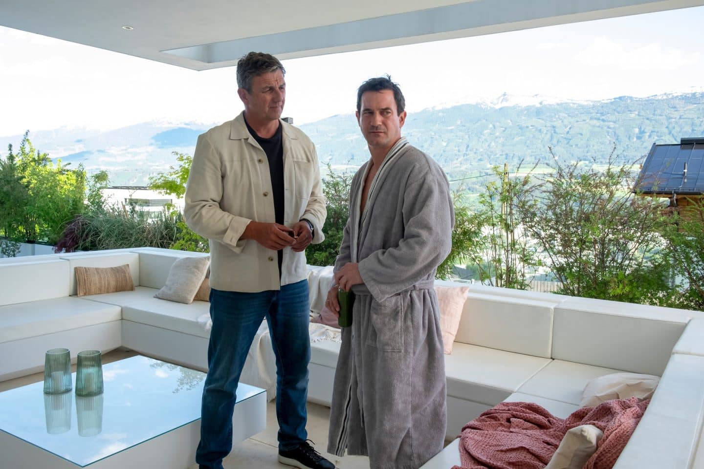 "Der Bergdoktor: Wechselwirkungen": Auf einer Terrasse mit Blick in die Berge: Dr. Martin Gruber (Hans Sigl) steht neben Hendrik Barlan (Sönke Möhring) und blickt ihn stirnrunzelnd an. Hendrik Barlan trägt einen Bademantel und hat sich mit nachdenklichem Blick von Dr. Gruber abgewendet.
