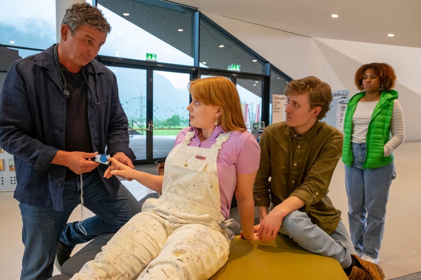 "Der Bergdoktor: Zwei Gesichter": Im Foyer des Festspielhauses: In ihrer Arbeitslatzhose sitzt Emily Scheffler (Lilly Charlotte Dreesen) auf einer Bank. Dr. Martin Gruber (Hans Sigl) steht neben ihr und misst mit einem Oximeter Lillys Puls. Hinter Lilly sitzt Oskar Lenz (Max Schimmelpfennig) und blickt Dr. Gruber fragend an. Im Hintergrund steht Sarah Meister (Joy Grant) und beobachtet das Geschehen mit besorgtem Blick.