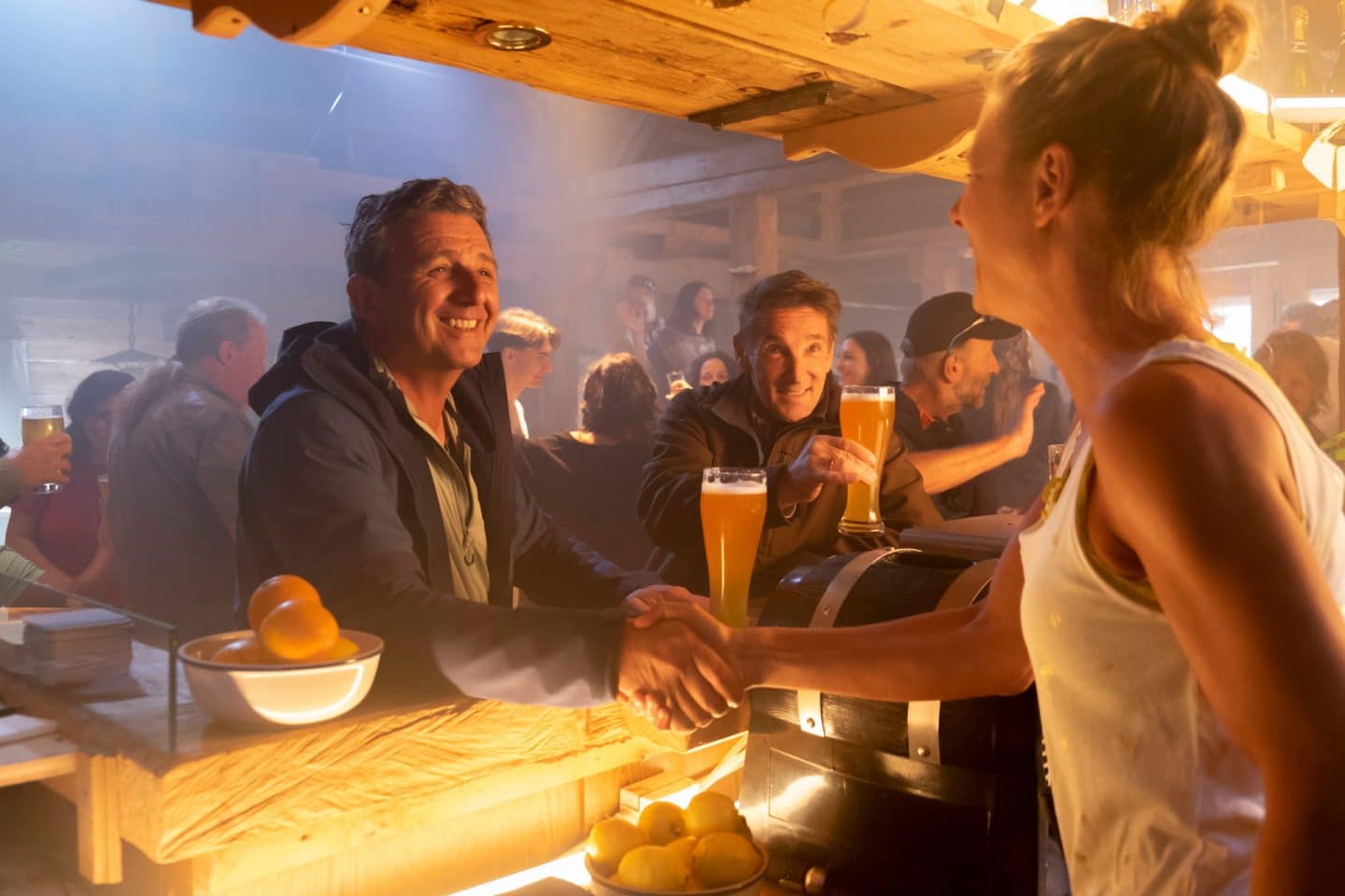 "Der Bergdoktor: Ein neuer Mensch": In der schummrig beleuchteten Funkenalm: Lächelnd reicht Martin Gruber (Hans Sigl) der Besitzerin Tina Reichert (Xenia Tiling) die Hand über den Tresen. Der neben ihm sitzende Alexander Kahnweiler (Mark Keller) prostet ihnen mit einem gefüllten Bierglas zu. Im Hintergrund tanzen Personen.
