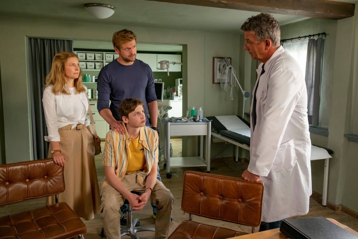 "Der Bergdoktor: Kindeswohl": Im Behandlungsraum der Arztpraxis: John Luckau (Nico Marischka) sitzt auf einem Hocker, seine Pflegeeltern Katharina Fischer (Birte Hanusrichter) und Benno Fischer (Steve Windolf) stehen hinter ihm. Besorgt blicken sie Dr. Martin Gruber (Hans Sigl) an, der vor ihnen steht.
