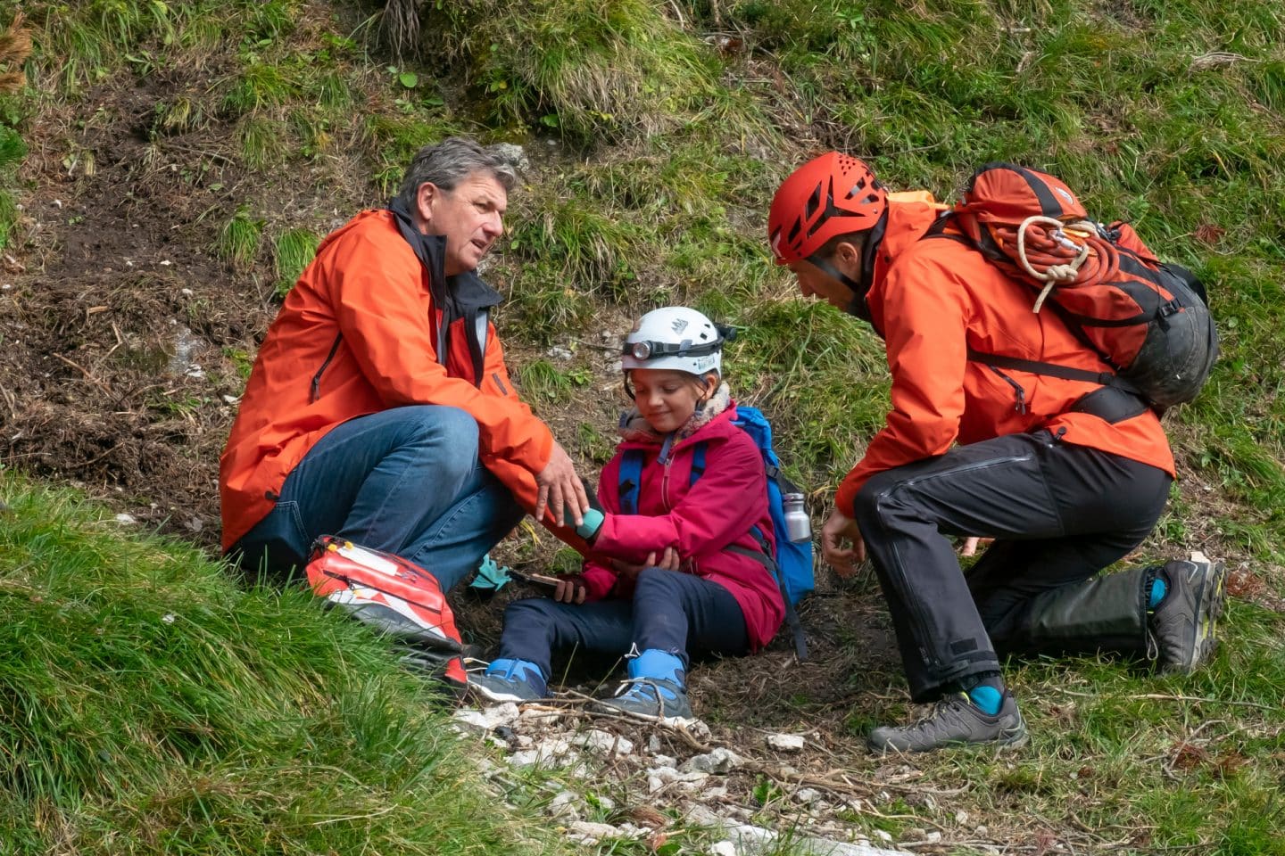 "Der Bergdoktor: Hoffnung": An einem begrünten Berghang: Tamara Gotthart (Rosa Wirtz) sitzt mit schmerzverzerrtem Gesicht am Boden. Sie trägt einen Helm mit Stirnlampe und einen Rucksack. Neben ihr sitzt Dr. Martin Gruber (Hans Sigl) und tastet ihren Arm ab, auf der anderen Seite kniet Hans Gruber (Heiko Ruprecht), der seine Bergretter-Ausrüstung trägt.
