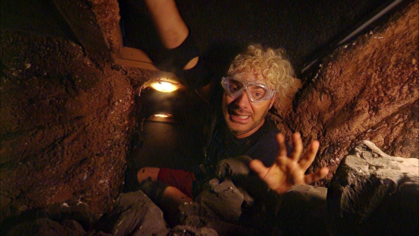 Sam Dylan kämpft sich durch die enge und dunkle Höhle der Dschungelprüfung 'Eruptions-Störung'. Mit Schutzbrille und Stirnlampe ausgestattet, versucht er, in dem felsigen, von Krabbeltieren bewohnten Tunnel die begehrten Sterne zu finden. Sein Gesichtsausdruck zeigt eine Mischung aus Konzentration und Angst, während er mit ausgestreckter Hand nach einem Hindernis tastet. Die beengte Umgebung, die feuchte Erde und die bedrohlich wirkende Beleuchtung verstärken die beklemmende Atmosphäre der Prüfung.