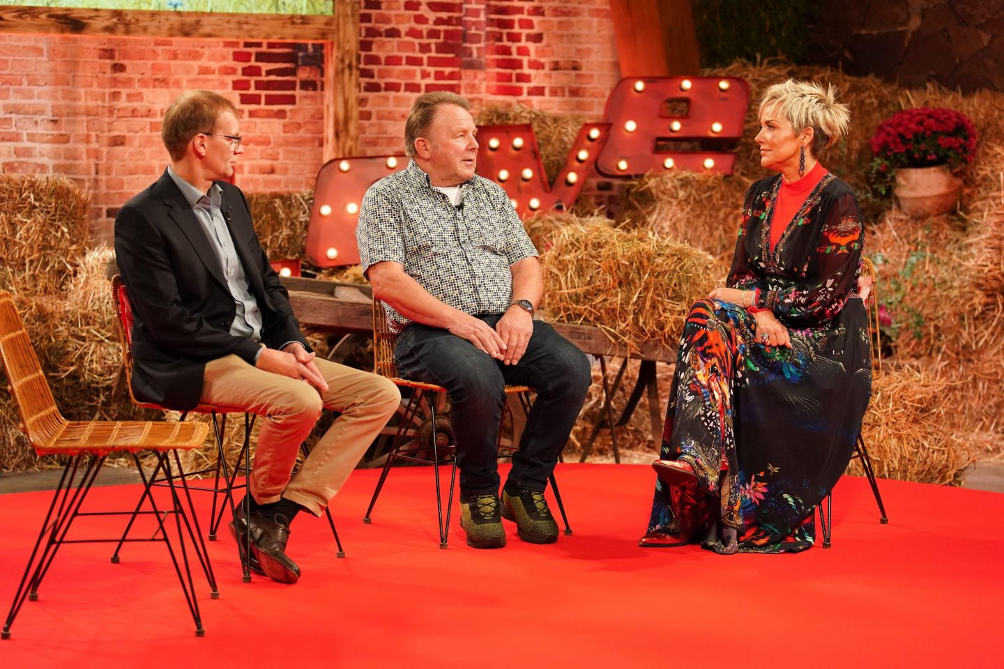 Olaf und Herbert sitzen auf Stühlen vor einer Kulisse aus Heuballen und sprechen mit Moderatorin Inka Bause, die ein farbenfrohes Kleid trägt. Die Szene findet auf einem roten Teppich statt, im Hintergrund leuchtet eine dekorative Schrift mit dem Wort „LOVE“.