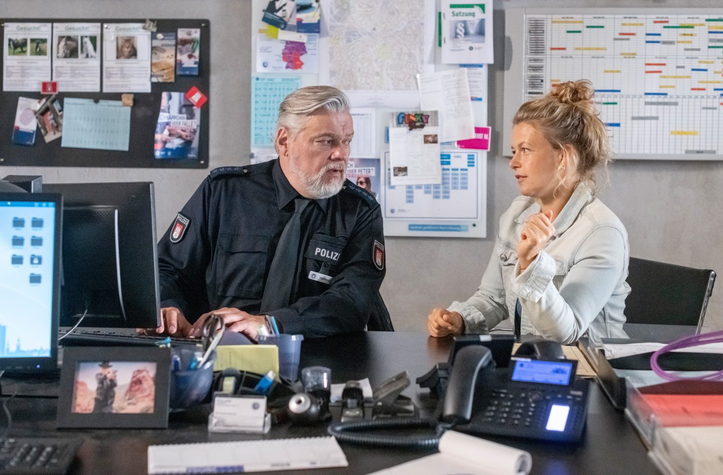 Helmut Husmann (Torsten Münchow, links) sitzt in Polizeiuniform an einem Schreibtisch und nimmt die Diebstahlsanzeige von Rebecca Sträther (Stella Burr, rechts) auf. Die Szene deutet auf eine Verbindung zu einem entführten Teenager-Mädchen hin.