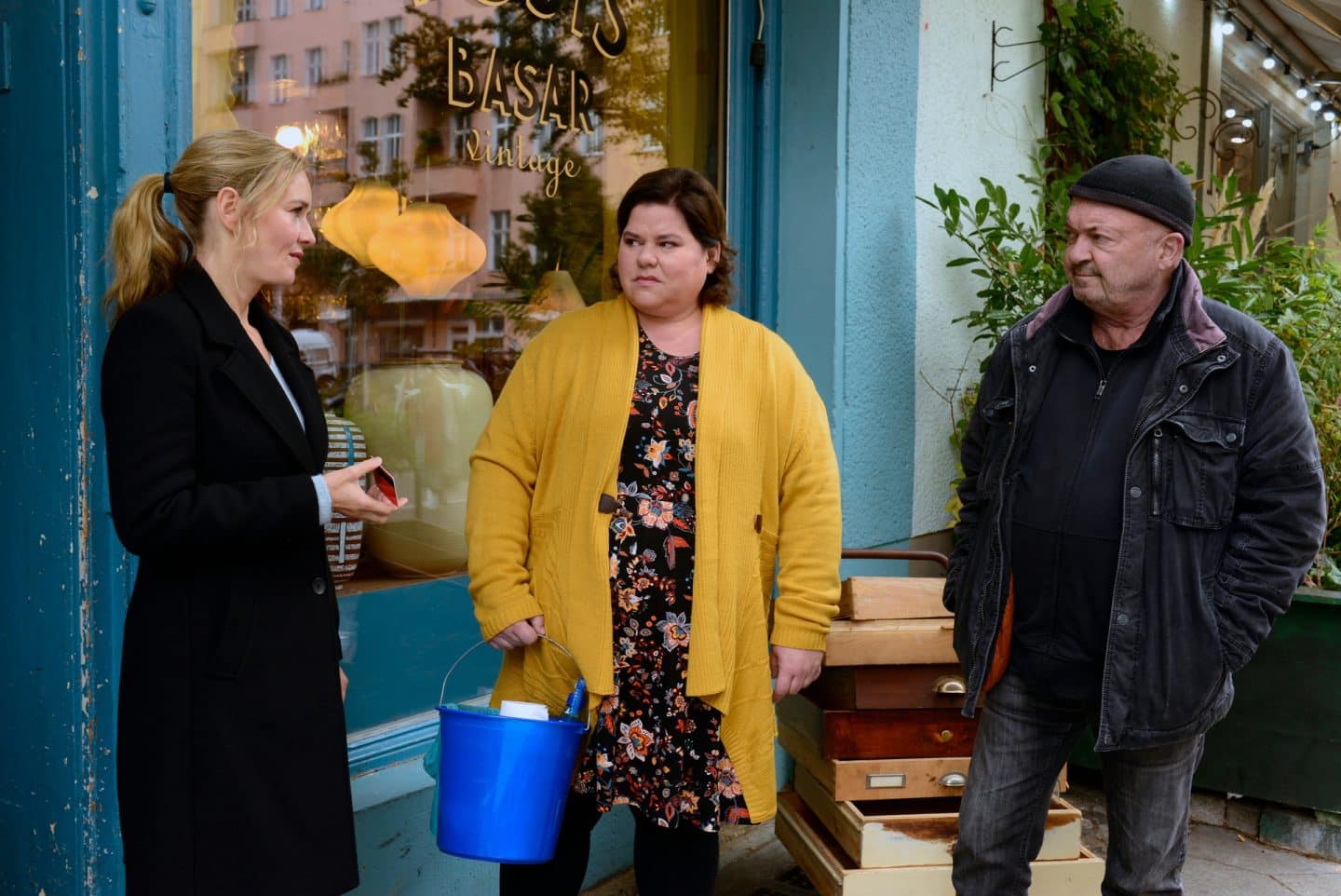 "Ein starkes Team - Der tote Mörder": Roswita Zander (Nadine Wrietz) steht vor ihrem Vintageladen und hält einen Eimer mit Putzmitteln in der Hand. Linett (Stefanie Stappenbeck) und Otto (Florian Martens) flankieren sie und werfen sich einen wissenden Blick zu. Roswita schaut Linett kritisch an, die ihr ihren Dienstausweis entgegenstreckt.
