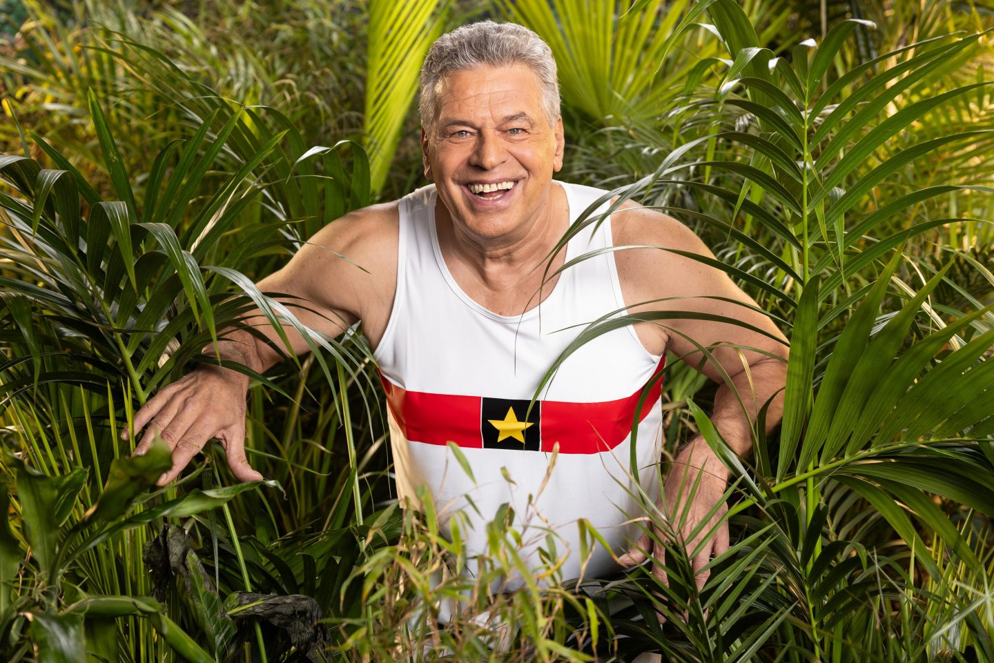 Jürgen Hingsen, ehemaliger deutscher Zehnkämpfer, in einem weißen Trikot mit rotem und schwarzem Streifen sowie gelbem Stern, lächelnd inmitten von grünem Dschungel.







