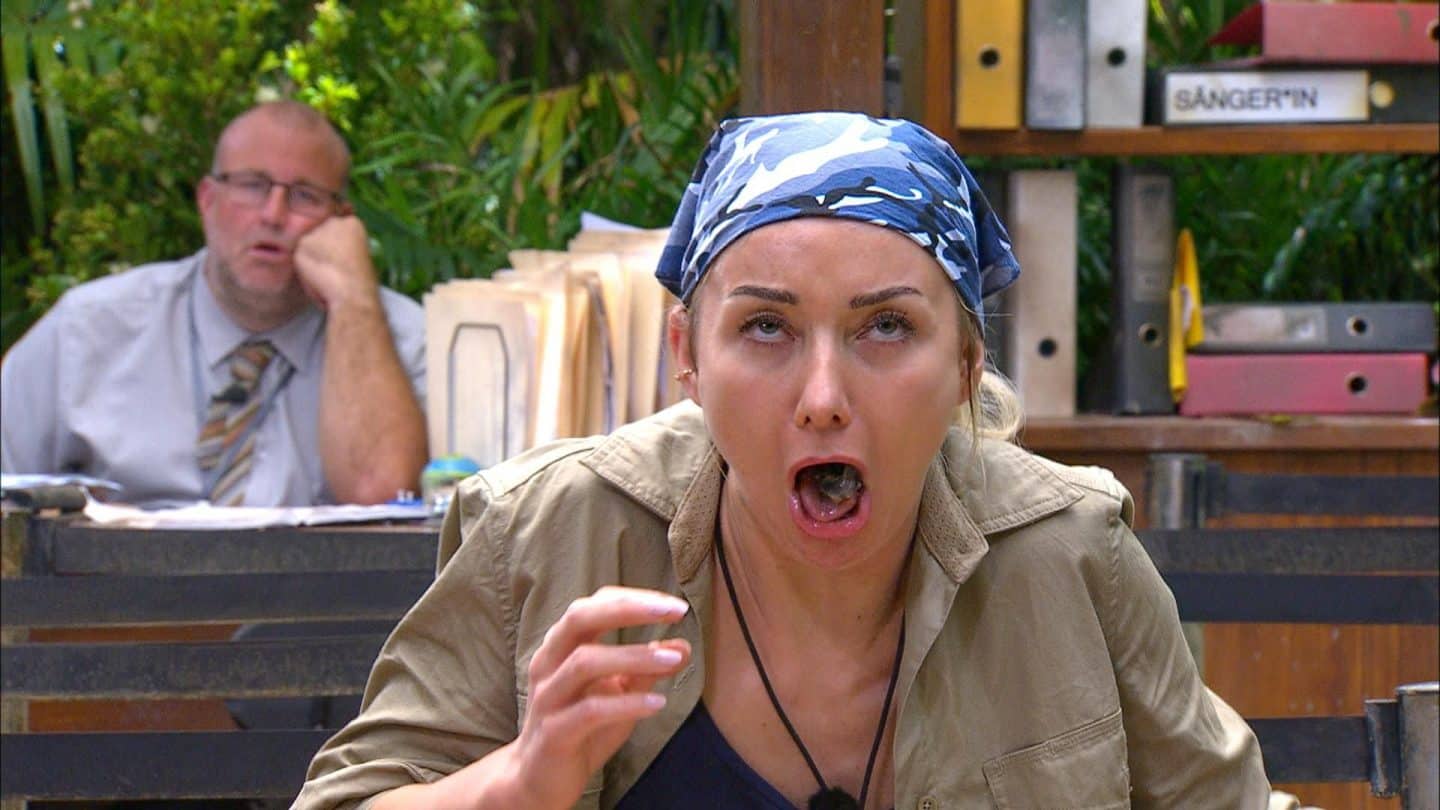 Anna-Carina Woitschack während der Dschungelprüfung „Bährufs-Bähratung“, trägt ein blau-weißes Kopftuch und eine beige Jacke, während sie mit offenem Mund kämpft, einen unappetitlichen Bissen herunterzuschlucken. Im Hintergrund sitzt ein Mann am Schreibtisch und beobachtet das Geschehen.