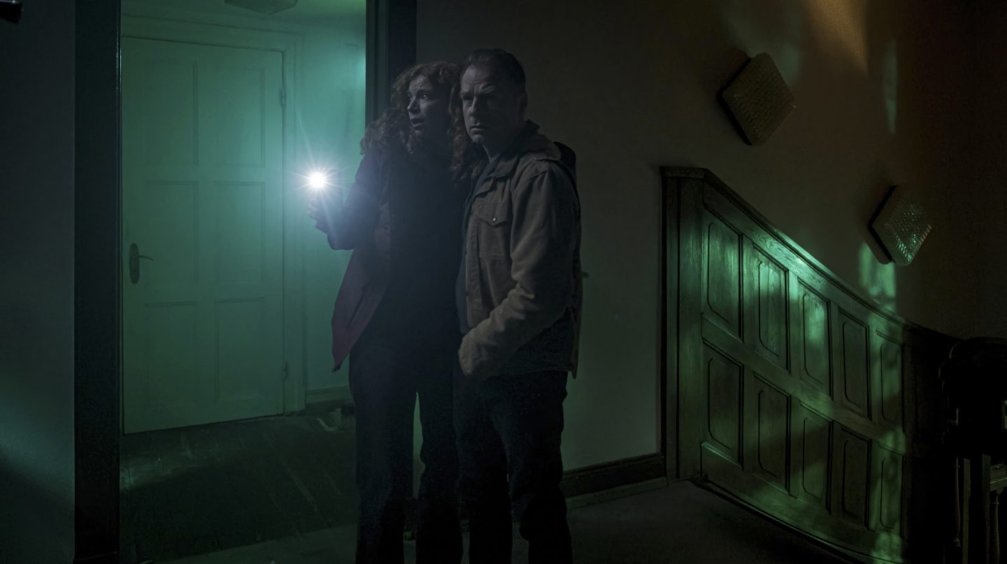 Jule Christiansen (gespielt von Marleen Lohse) und Hauke Jacobs (gespielt von Hinnerk Schönemann) stehen mit einer Taschenlampe in einem düsteren Treppenhaus. Die Szenerie ist von unheimlichem grünlichem Licht durchzogen, was die angespannte Atmosphäre im Nolden-Haus unterstreicht.