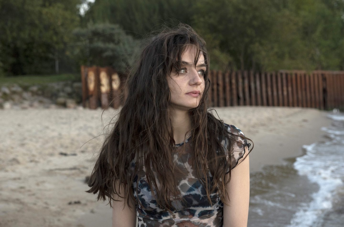 Nele Krüger (Laura Balzer), eine junge Frau mit langen, zerzausten Haaren steht nachdenklich am Strand. Im Hintergrund sind Sand, eine Uferböschung mit Vegetation und eine rostige Metallwand zu sehen. Die Szene vermittelt eine melancholische und geheimnisvolle Atmosphäre.