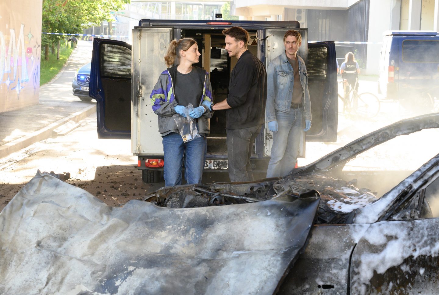 Nach dem nächtlichen Überfall auf einen Geldtransporter stehen die Ermittler am Tatort. Hauptkommissarin Pia Heinrich (Ines Marie Westernströer) trägt Handschuhe und hält eine durchsichtige Beweismitteltüte in den Händen, während sie sich mit Hauptkommissar Leo Hölzer (Vladimir Burlakov) unterhält, der in schwarzer Jacke neben ihr steht. Im Hintergrund lehnt Hauptkommissar Adam Schürk (Daniel Sträßer) in Jeansjacke mit ernstem Blick an einem geöffneten Einsatzfahrzeug. Ein ausgebranntes Autowrack ist im Vordergrund zu sehen, während eine Polizistin im Hintergrund an einem Absperrband steht. Rauch und Schaumreste sind auf der Szene verteilt.