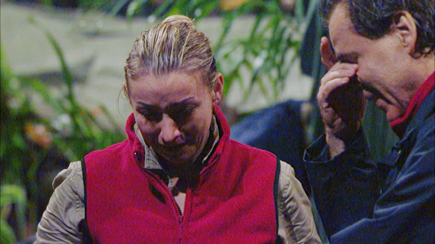 Anna-Carina Woitschack und Jörg Dahlmann stehen im Dschungelcamp, sichtlich bewegt von einem emotionalen Gespräch. Anna-Carina trägt eine rote Weste über einer beigen Bluse und senkt den Blick, während Tränen in ihren Augen stehen. Jörg, ebenfalls tief betroffen, reibt sich die Stirn mit einer Hand und wirkt nachdenklich. Die dichte, grüne Vegetation im Hintergrund verstärkt die nachdenkliche und bedrückte Atmosphäre. Das Thema ihrer Unterhaltung: die schmerzhafte Trennung von Stefan Mross, die für die Sängerin eine schwere Zeit bedeutet hat.