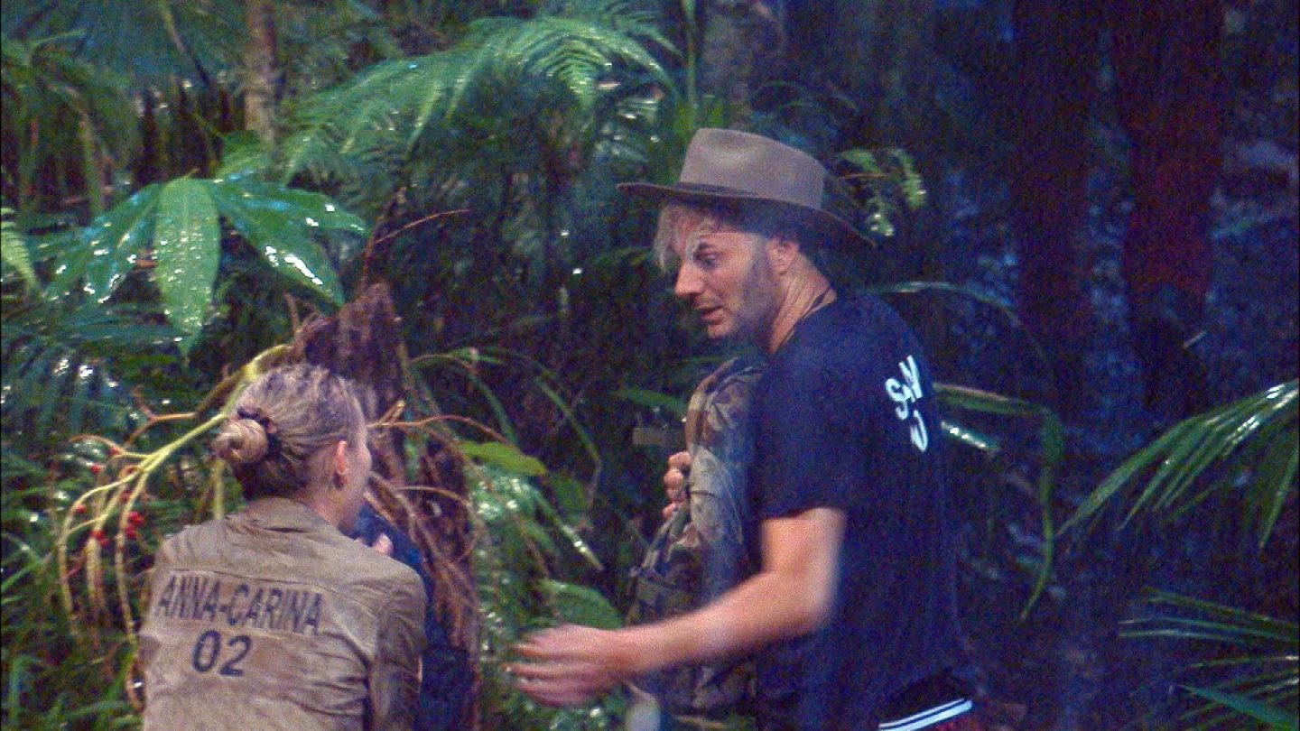 Anna-Carina Woitschack und Sam Dylan flüchten durch den strömenden Regen im dichten Dschungel. Anna-Carina trägt eine durchnässte khakifarbene Jacke mit ihrem Namen und der Nummer '02' auf dem Rücken. Ihr nasses Haar ist zu einem Knoten gebunden. Sam Dylan, mit einem breiten Hut und einem dunklen T-Shirt, hält seinen Rucksack fest und blickt mit angespannter Miene zurück. Der dichte Regenwald mit tropischen Pflanzen bildet den Hintergrund, während die beiden nach Schutz suchen.