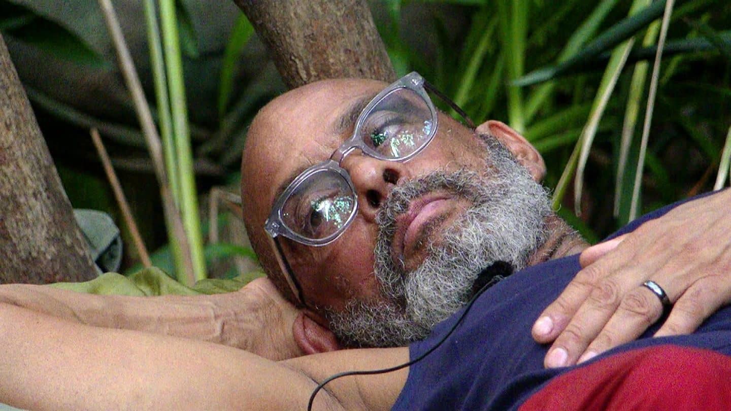 Pierre Sanoussi-Bliss liegt entspannt im Dschungelcamp auf einer Unterlage und lehnt seinen Kopf an einen Baumstamm. Mit nachdenklichem Blick und durchsichtiger Brille scheint er in die Ferne zu schauen.