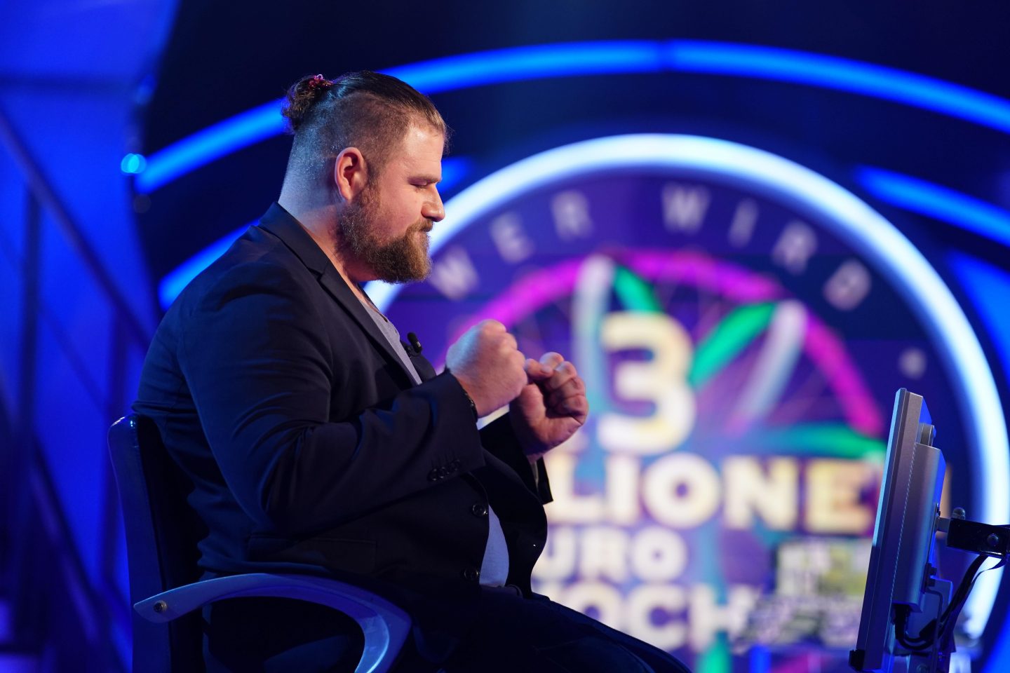 Ein Mann mit Bart und zusammengebundenem Haar sitzt auf einem Stuhl in einem TV-Studio, hebt die Fäuste in einer Jubelgeste und lächelt. Im Hintergrund ist das Logo der „3-Millionen-Euro-Woche“ von „Wer wird Millionär?“ zu sehen.






