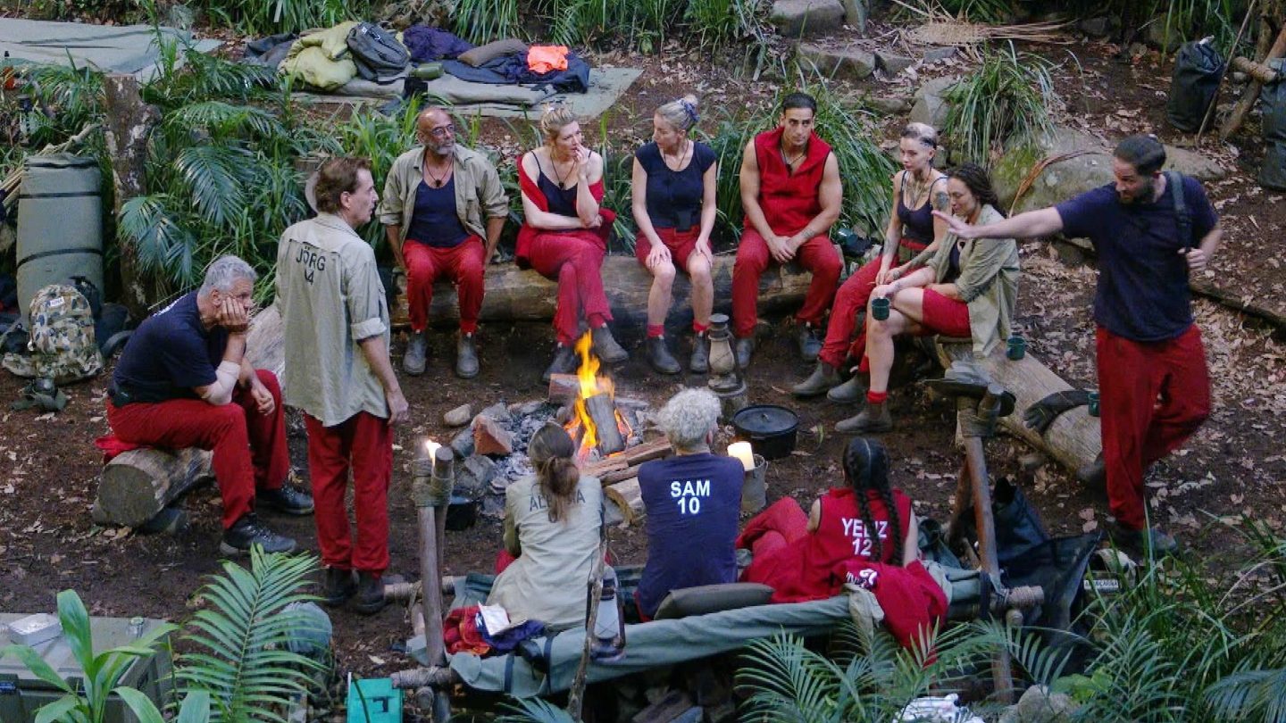 Die Dschungelcamp-Bewohner sitzen in einem Kreis um ein Lagerfeuer, während Maurice Dziwak (rechts) aufsteht, seinen Rucksack schultert und gestikulierend in Richtung Dschungeltelefon geht. Die übrigen Camper, darunter Jörg, Alessia, Sam und Yeliz, beobachten die Szene – einige nachdenklich, andere sichtlich erschöpft. Im Hintergrund sind provisorische Schlafplätze und Ausrüstungsgegenstände zu sehen.
