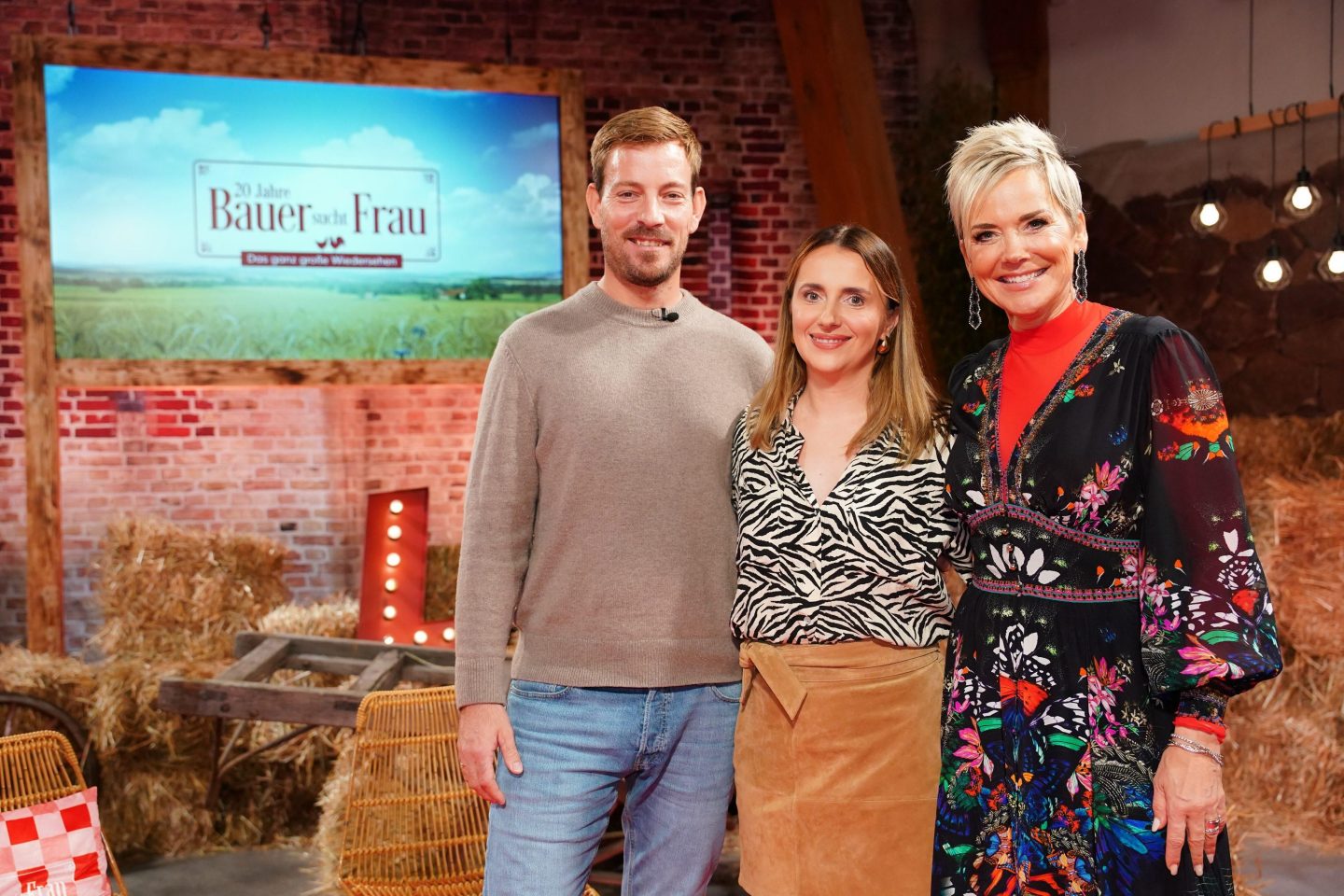 Gerald und Anna stehen neben Moderatorin Inka Bause im rustikal eingerichteten Studio von '20 Jahre Bauer sucht Frau – Das ganz große Wiedersehen'. Gerald trägt einen grauen Pullover und blaue Jeans, Anna ein schwarz-weiß gemustertes Oberteil und einen braunen Rock. Inka Bause strahlt in einem bunten Kleid. Im Hintergrund ist eine rustikale Kulisse mit Heuballen und dekorativen Lichtern zu sehen.