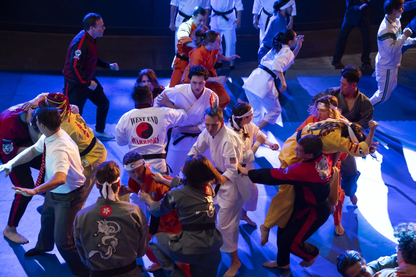 Kampfszene in einer großen Halle: Mehrere Karate-Kämpfer in verschiedenen Kampfsportuniformen liefern sich einen intensiven Massenkampf. In der Mitte steht ein junger Mann in einem weißen Karate-Gi, während um ihn herum Kämpfer mit bunten Uniformen und Stirnbändern in dynamischen Bewegungen verwickelt sind. Die Szene ist dramatisch beleuchtet, mit blauen und violetten Lichtakzenten.