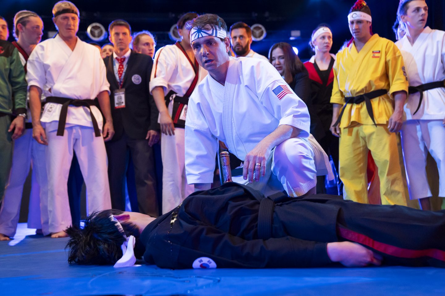 Daniel LaRusso (Ralph Macchio) in einem weißen Karate-Gi mit USA-Flaggenaufnäher kniet mit besorgtem Gesichtsausdruck neben einem bewusstlosen Kämpfer in schwarzer Kampfsportkleidung auf einer blauen Matte. Um sie herum stehen andere Kämpfer und Kampfrichter in verschiedenen Karate-Uniformen, die mit ernsten Mienen auf die Szene blicken. Im Hintergrund sorgt blaues Licht für eine dramatische Atmosphäre.