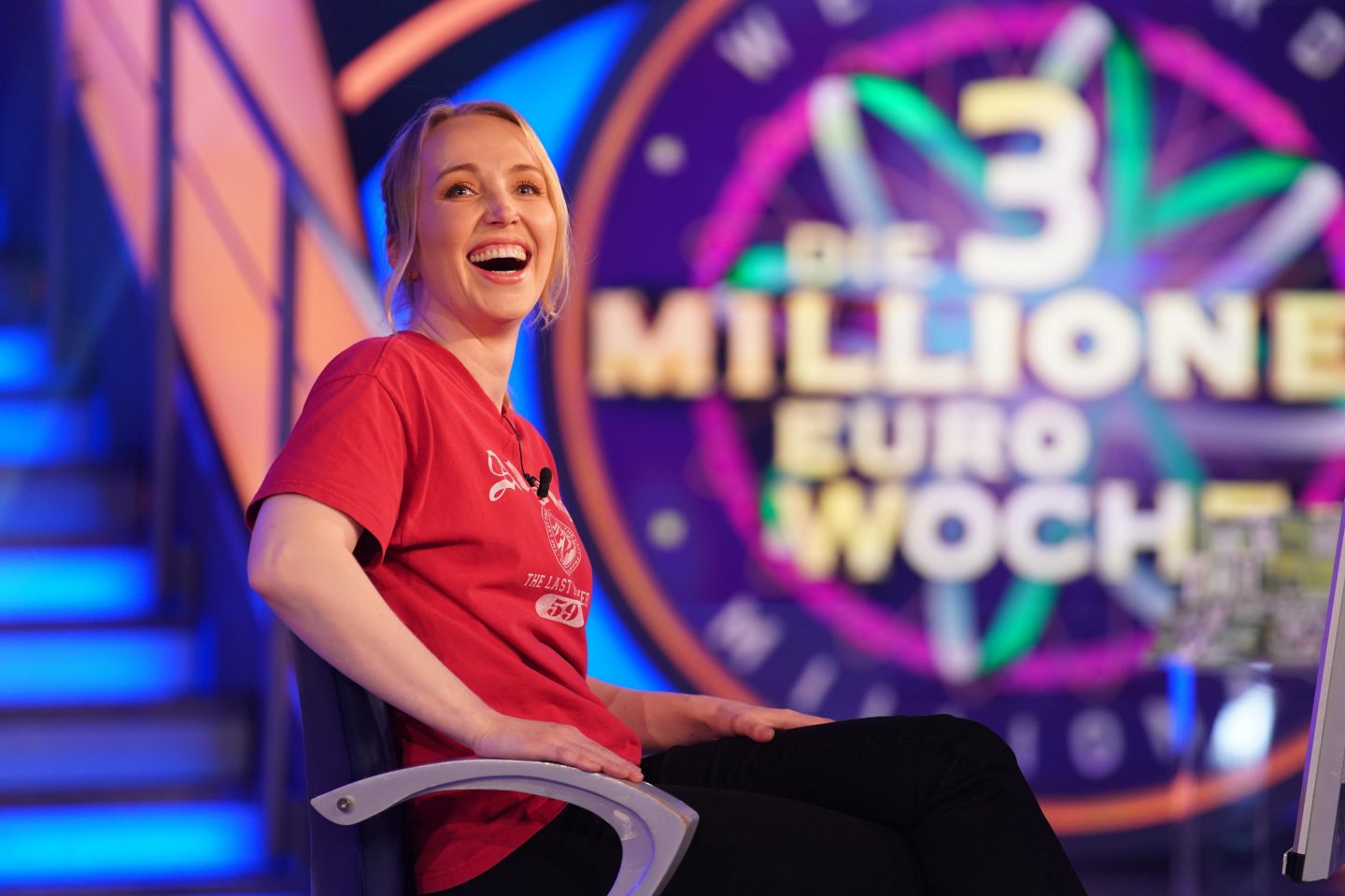Dr. Paula Menold, eine blonde Frau im roten T-Shirt, sitzt lächelnd auf einem Stuhl im TV-Studio. Im Hintergrund ist das Logo der „3-Millionen-Euro-Woche“ von „Wer wird Millionär?“ zu erkennen.






