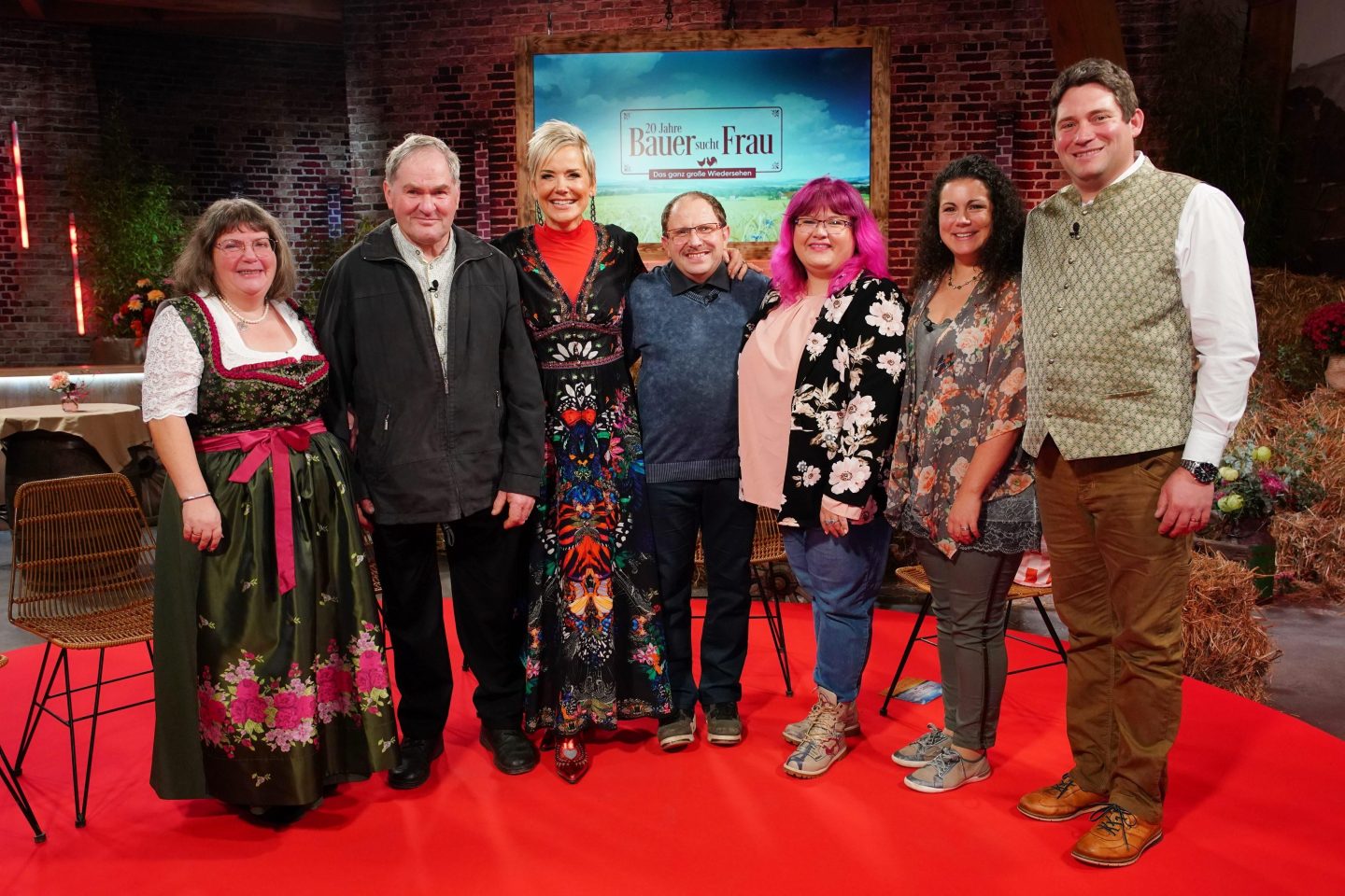 v.l.) Sabine und Albert, Moderatorin Inka Bause, Steffen und Nicole, Victoria und Guy stehen lächelnd nebeneinander auf einem roten Teppich vor der festlichen Kulisse der Jubiläumsshow „20 Jahre Bauer sucht Frau“. Im Hintergrund sind Heuballen, Blumenarrangements und das Logo der Sendung zu sehen.