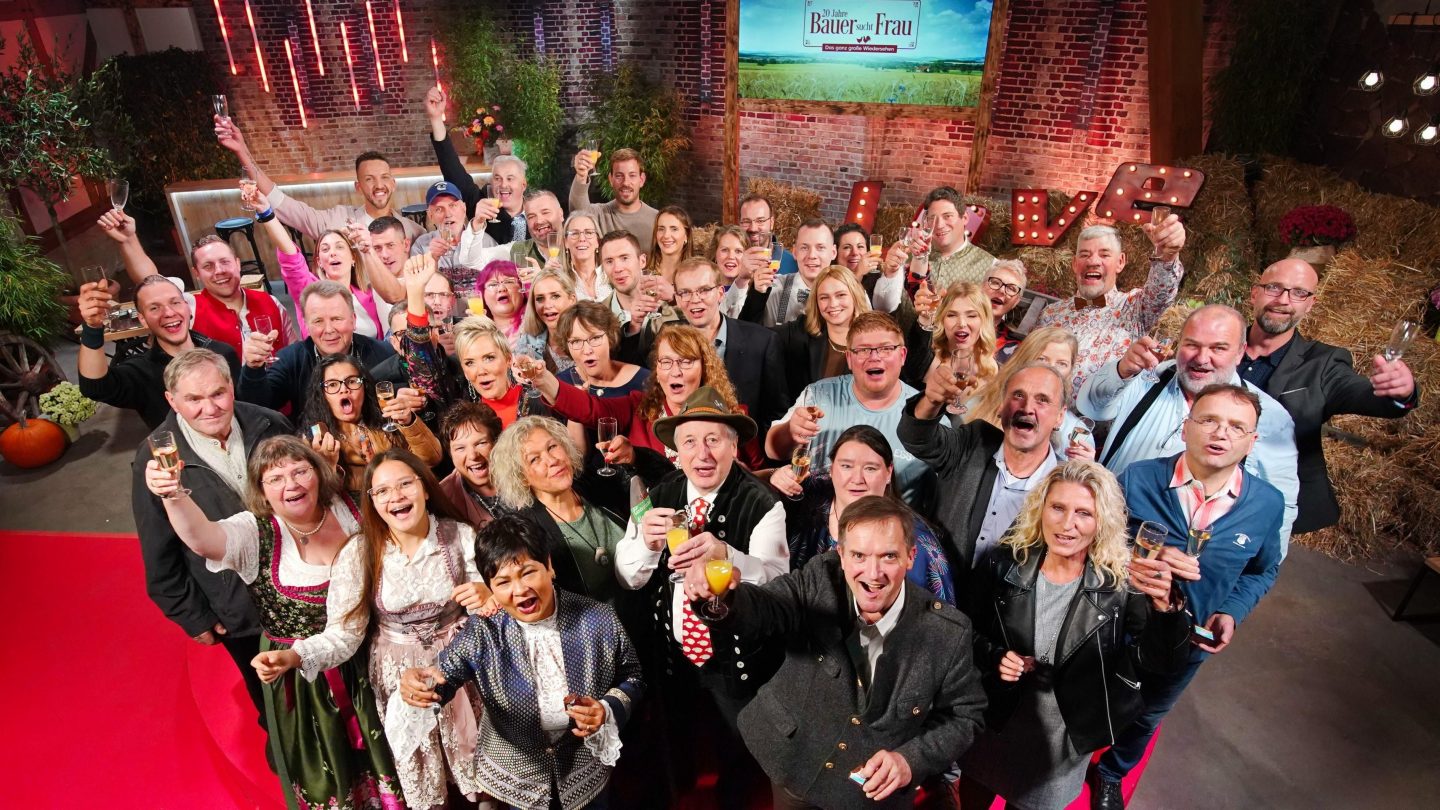 Gruppenfoto mit Teilnehmern aus 20 Jahren „Bauer sucht Frau“. Männer und Frauen stoßen fröhlich mit Gläsern an, um das Jubiläum der beliebten TV-Sendung zu feiern. Im Hintergrund ist eine festliche Kulisse mit Heuballen, einem roten Teppich und einer Leuchtschrift „LOVE“ zu sehen.