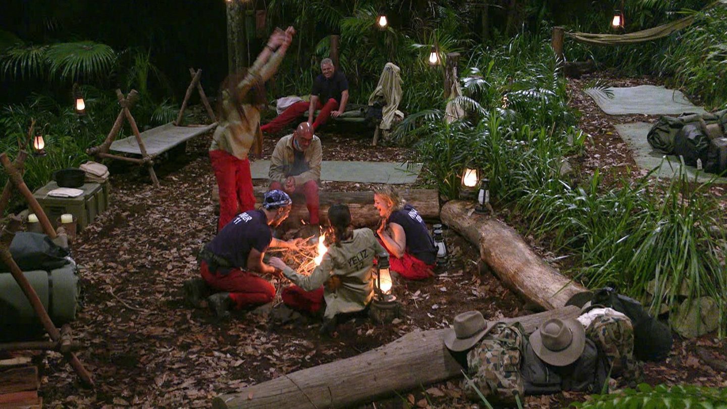 Dschungelcamp-Bewohner Lilly Becker, Timur Ülker, Pierre Sanoussi-Bliss, Jürgen Hingsen, Yeliz Koç und Nina Bott sitzen im Kreis um ein Lagerfeuer, während sie sich über das Entzünden des Feuers freuen. Im Hintergrund sind Hängematten und Campingausrüstung zu sehen.