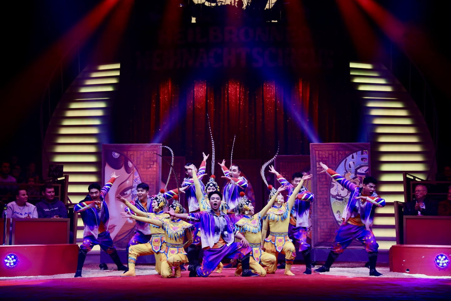 Die Dezhou Acrobatic Gruppe führt eine beeindruckende Darbietung auf einer Zirkusbühne auf. Die Künstler tragen bunte, traditionelle Kostüme in Gelb, Blau und Lila mit aufwendigen Verzierungen. Sie posieren in einer dynamischen Formation, einige mit erhobenen Armen, während Federschmuck ihre Bewegungen betont. Im Hintergrund ist eine festlich beleuchtete Bühne mit roten Vorhängen und dekorativen Elementen zu sehen. Zuschauer applaudieren am Rand der Manege.






