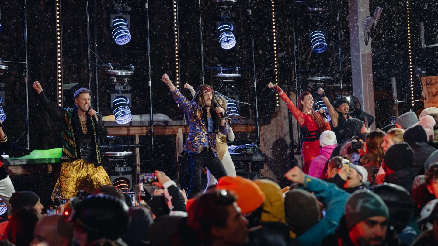 Schlager-Party beim MooserWirt in St. Anton: Künstler in bunten Outfits heizen der Menge ein, während Schneeflocken in der Luft tanzen. Das Publikum feiert ausgelassen vor der Bühne mit hochgereckten Armen und Smartphones in der Hand