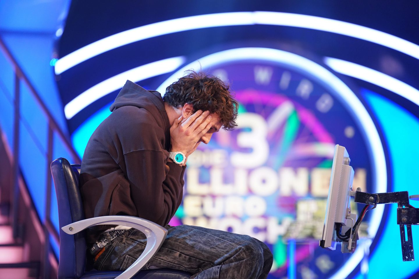 Junger Mann in braunem Hoodie, Kopf in den Händen, sitzt verzweifelt auf einem Stuhl vor dem Logo der „3-Millionen-Euro-Woche“ in der Quizshow „Wer wird Millionär?“.







