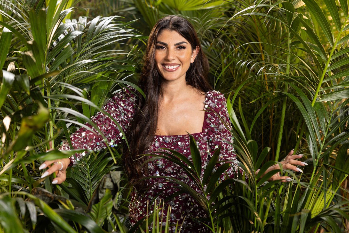 Yeliz Koc, lächelnd in einem bordeauxfarbenen Kleid mit glitzernden Details, mit langen dunklen Haaren, umgeben von tropischen Pflanzen im Dschungel.







