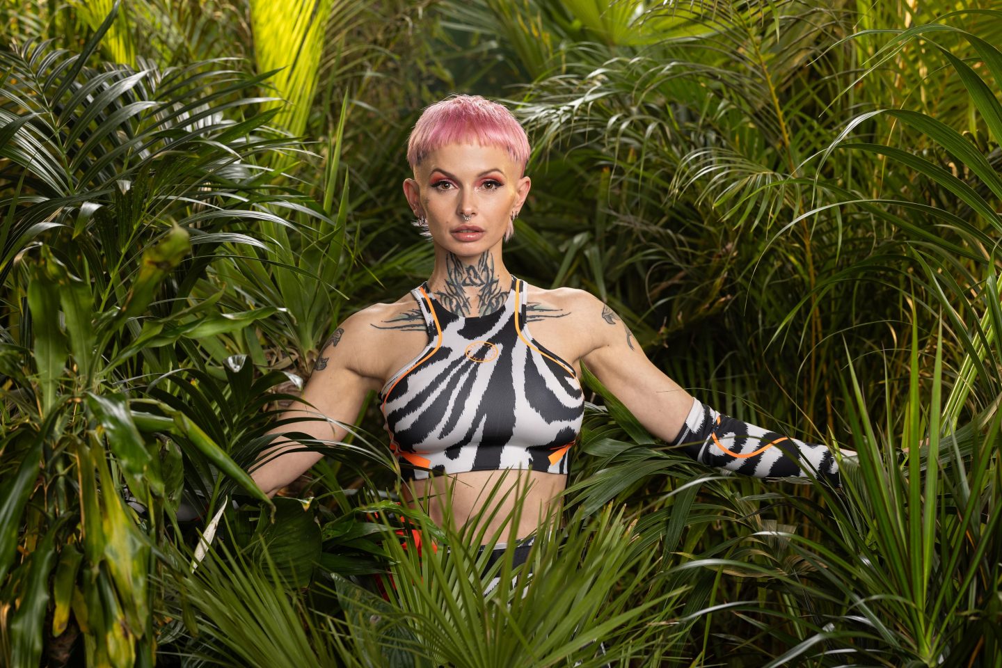 Edith Stehfest, in einem zebraähnlichen Sporttop mit auffälligen Tattoos am Hals und an den Armen, mit kurzen pinkfarbenen Haaren, umgeben von tropischen Pflanzen im Dschungel.







