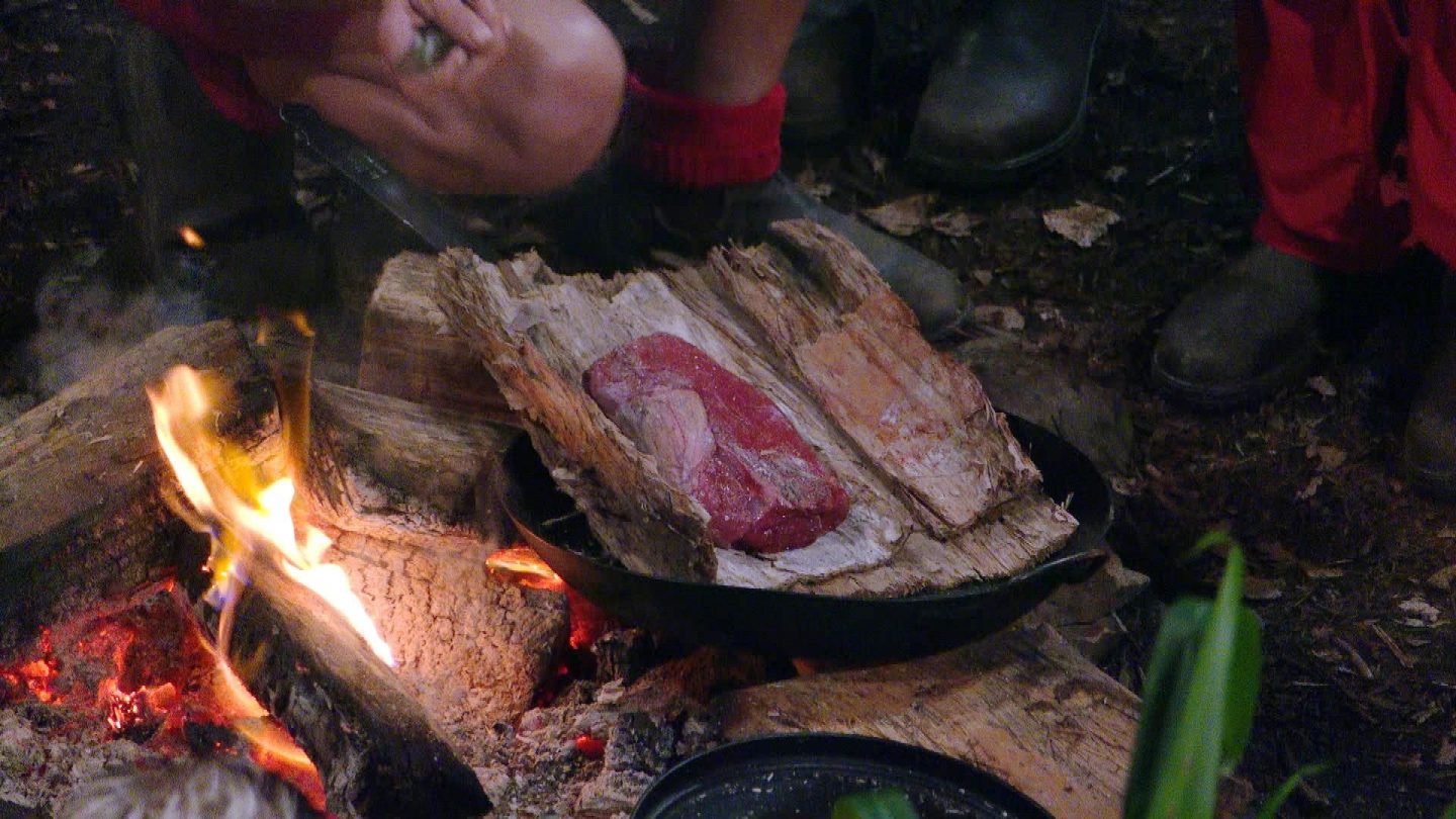 Ein Stück rohes Büffelnackenfleisch liegt auf einem Stück Holz über einer Pfanne am offenen Lagerfeuer im Dschungelcamp. Die Flammen lodern, während ein Teilnehmer das Fleisch mit einem Messer vorbereitet. Rundherum sind weitere Zutaten wie Süßkartoffeln und Paprika zu sehen.